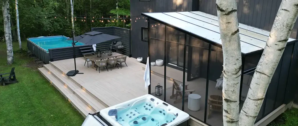 Backside of a modern home with a screened-in outdoor sitting area features a light tan deck with an above ground pool and hot tub.