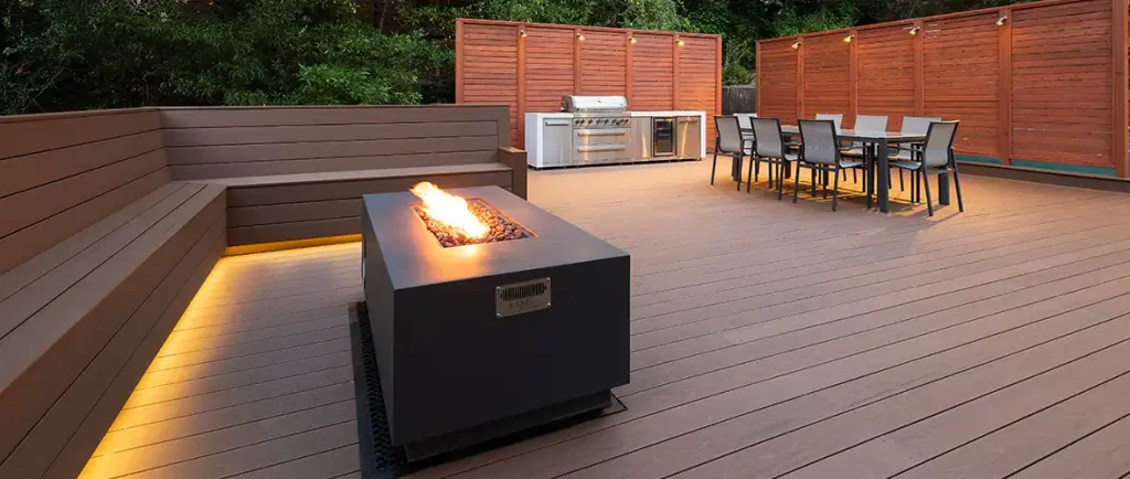 Dark brown deck with built-in benches around a modern free-standing fireplace. Lighting has been integrated into the underside of the benches.