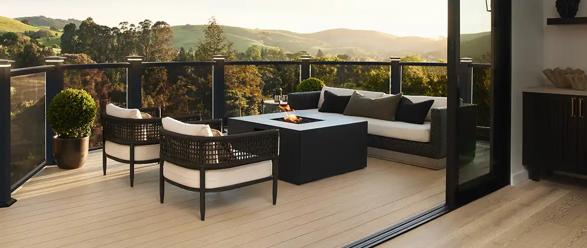 Tan deck with black railing and glass infill. An outdoor fireplace with furniture surrounding it is on the deck and rolling hills are visible in the background