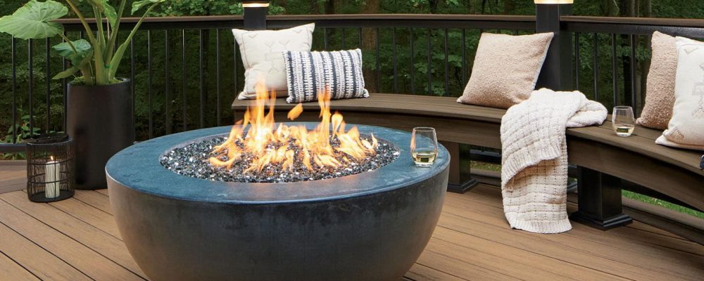 A round fire pit on a wooden deck with a curved bench and pillows, surrounded by a forest background