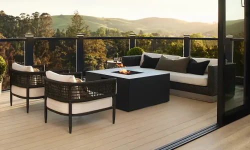 Tan deck with black railing and glass infill. An outdoor fireplace with furniture surrounding it is on the deck and rolling hills are visible in the background